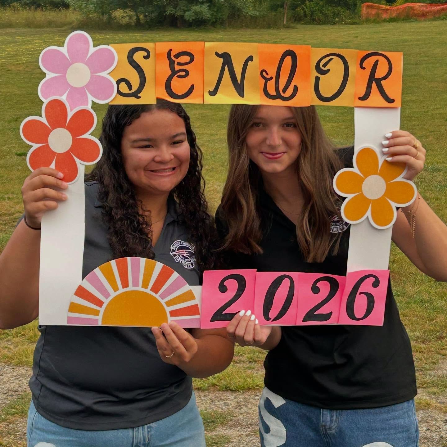 Two seniors holding a hand-lettered Senior 2026 photo frame on the campus lawn