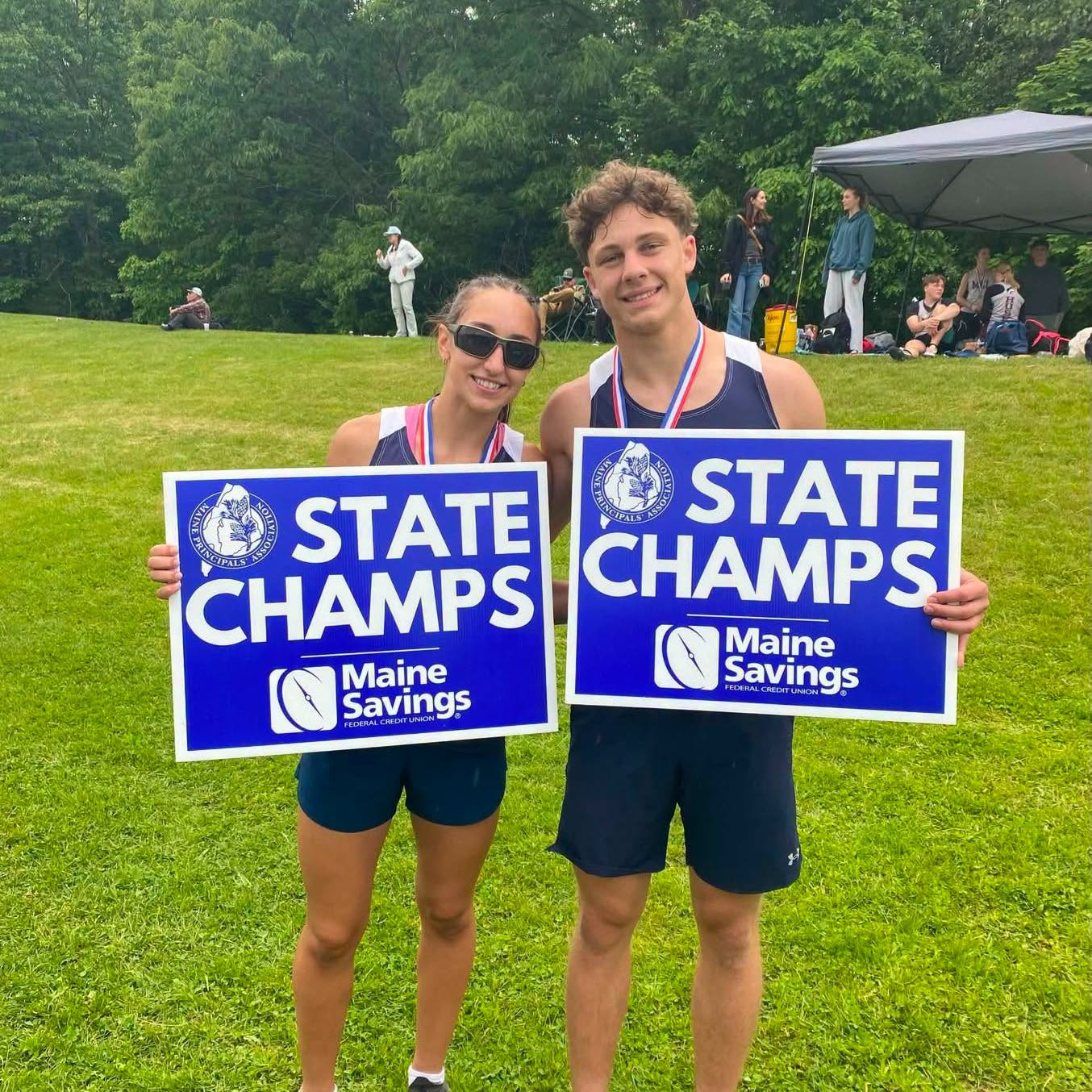 Two Patriots track athletes holding State Champs signs with medals around their necks